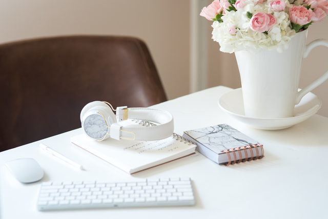 A white table surface styled with a keyboard, flowers in a vase, notebooks, headphones, and a computer mouse.
